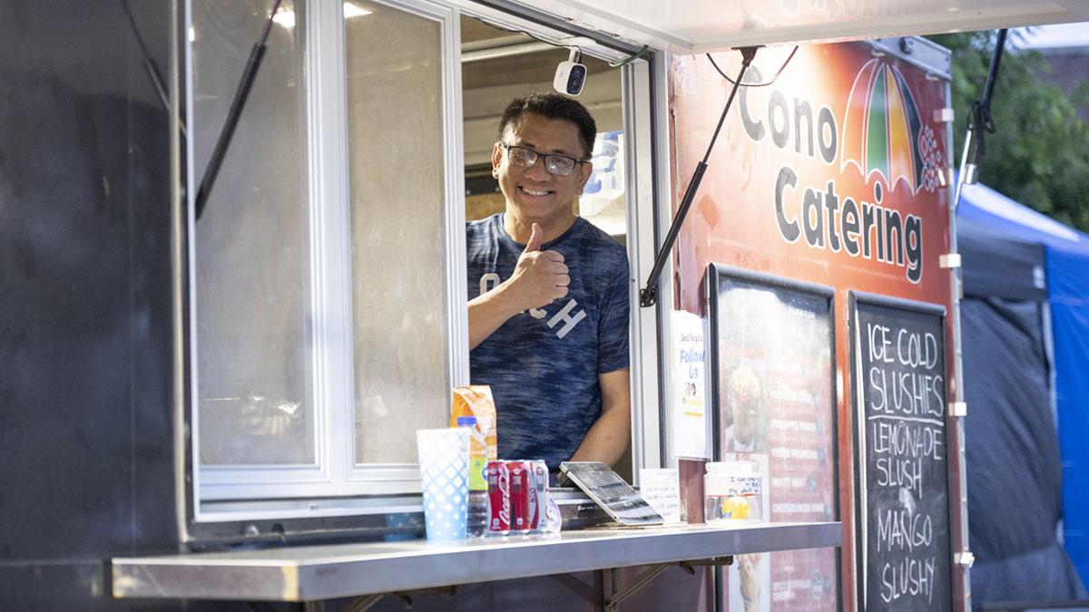 A smiling vendor gives a thumbs-up from a food truck window.