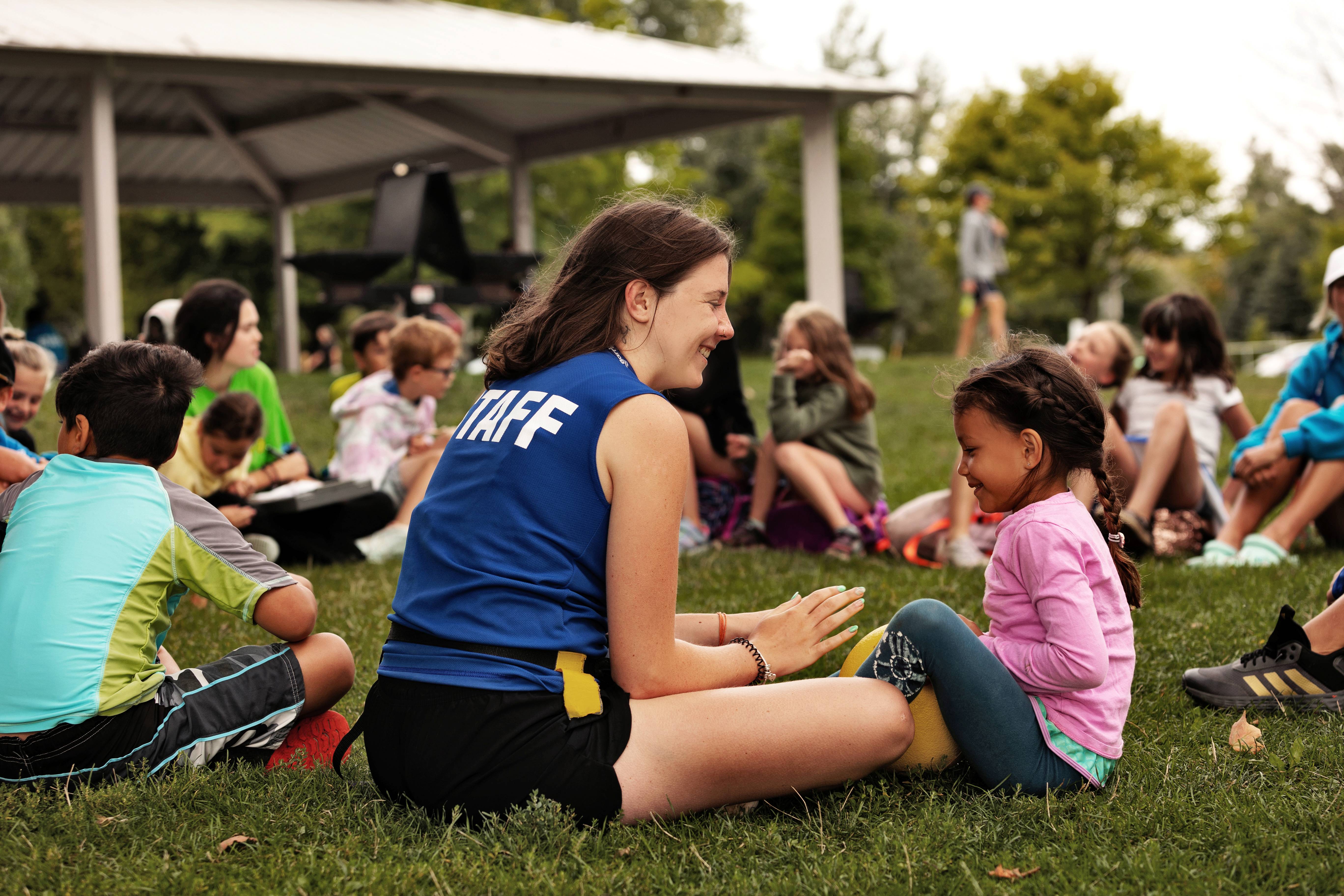 Rec staff person talking with child, sitting on grass