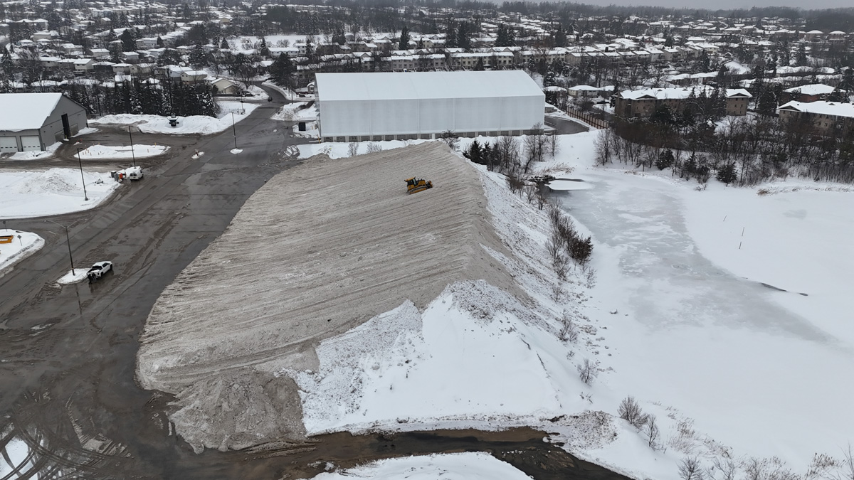 Aerial view of a large outdoor snow storage facility in winter, with a large pile of snow and surrounding buildings.
