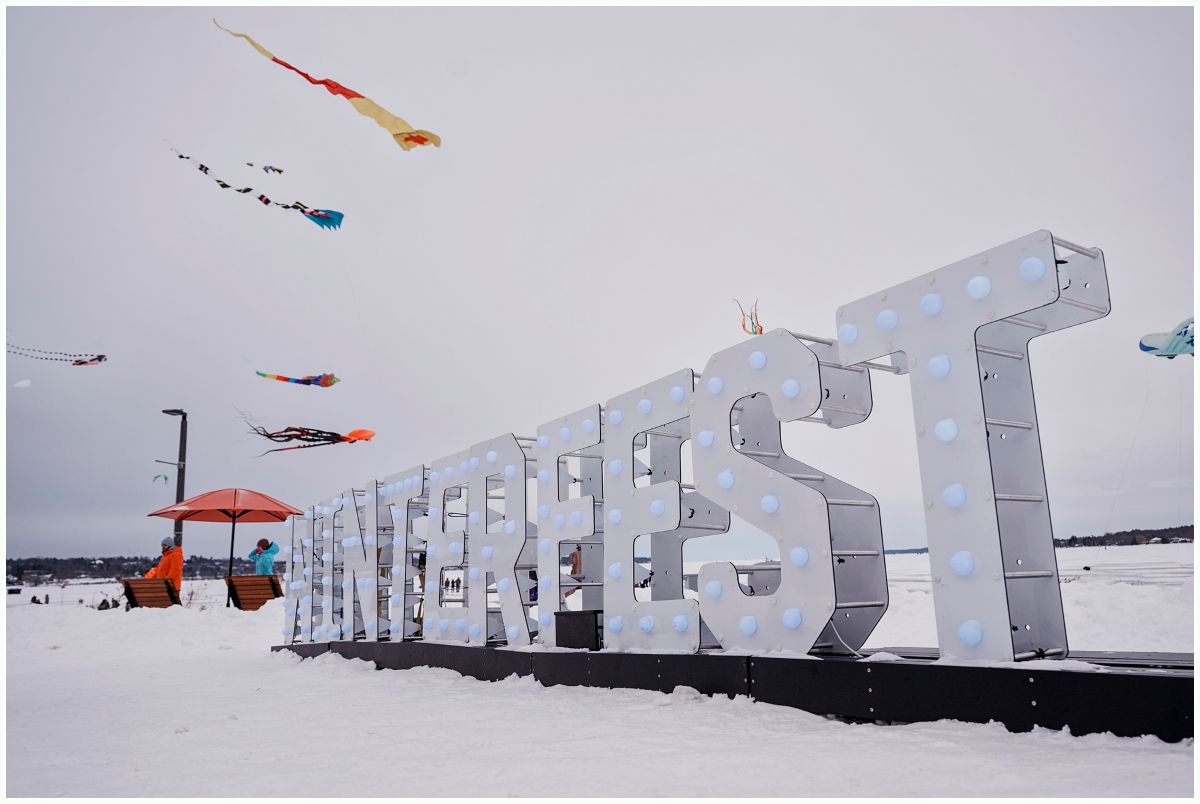 Kites in the sky over a large Winterfest sign, surrounded by snow. 