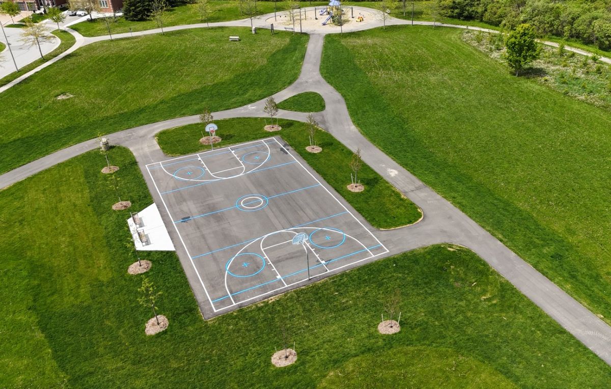 Aerial view of a multi-use court surrounded by newly planted trees and green space, with a playground in the background.