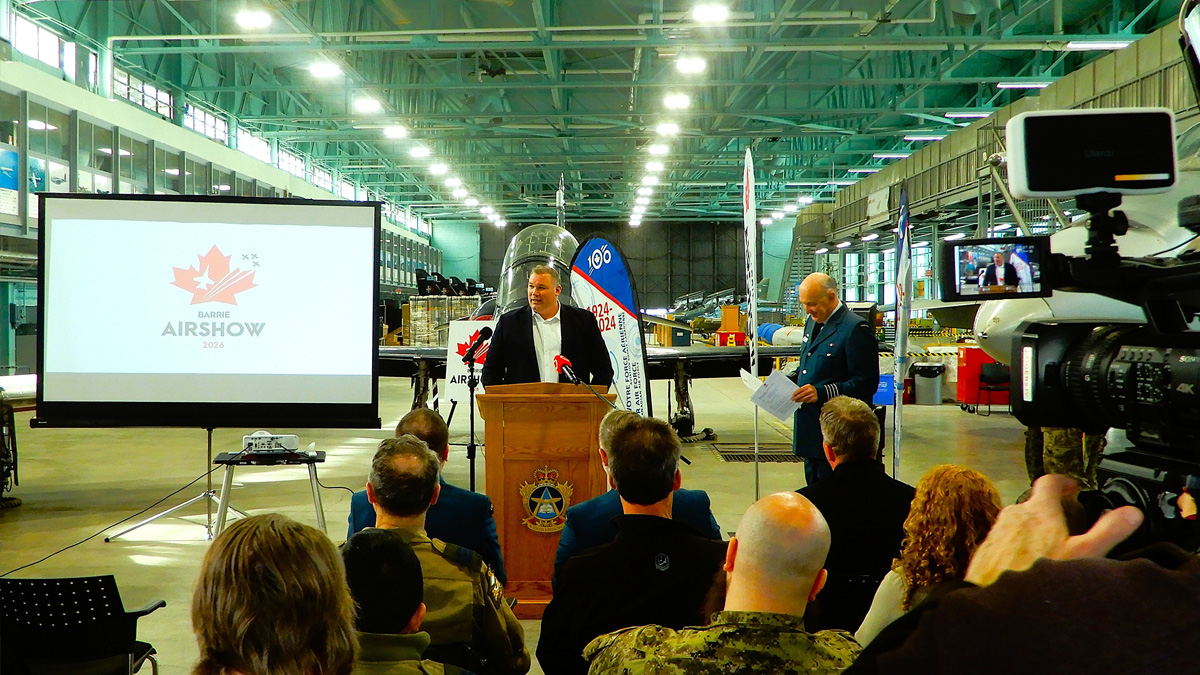 Speaker at podium in aircraft hangar with audience and camera crew.