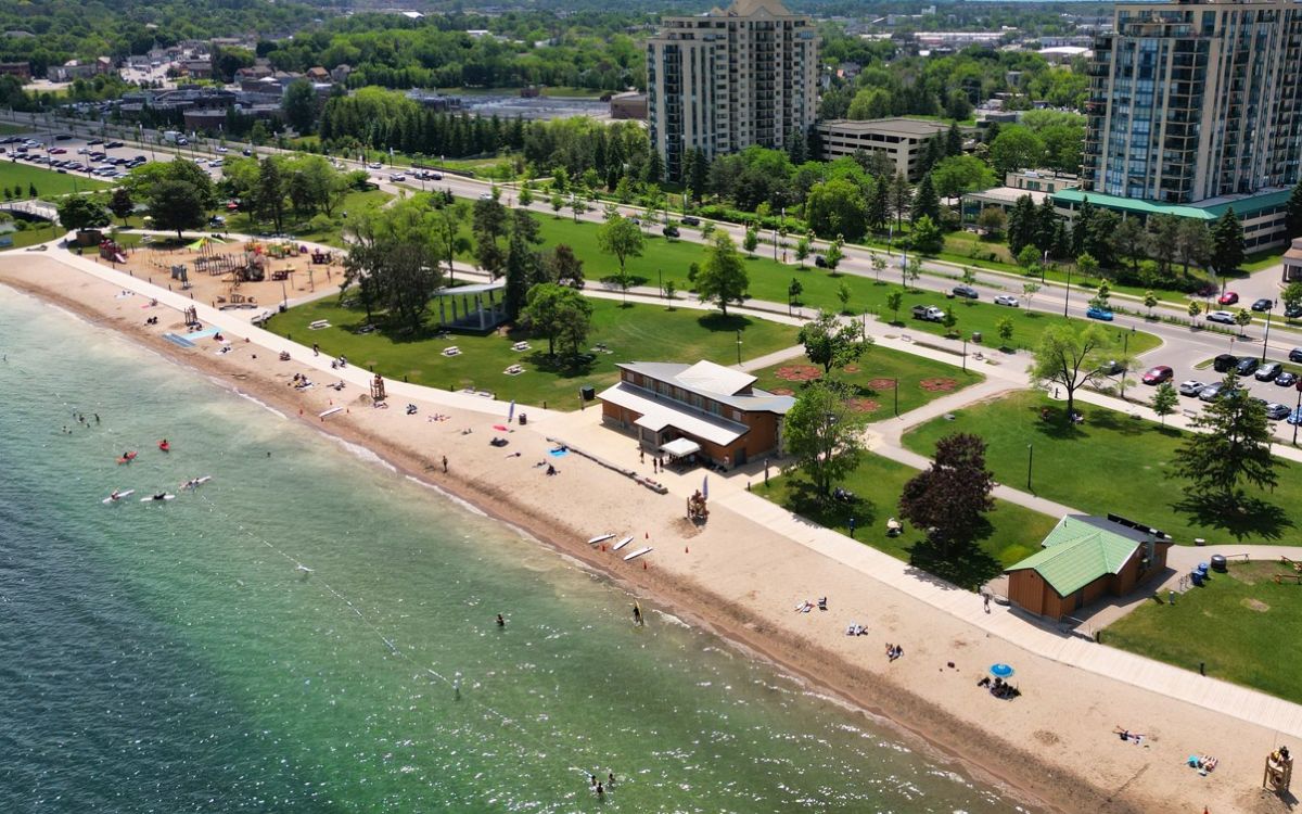 Aerial view of a beach with people, sandy shore, green parkland, and tall buildings in the background.