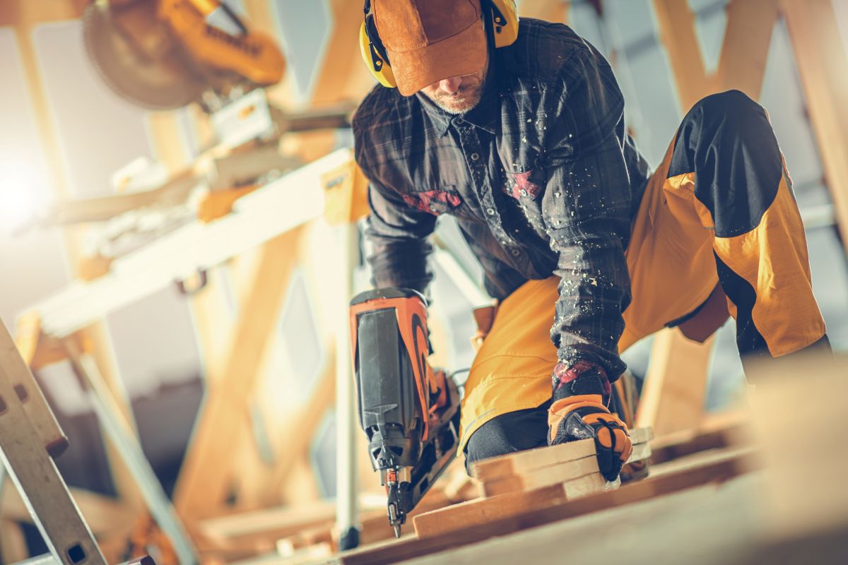 A construction worker kneels on a wooden floor using a nail gun in a bright workshop.