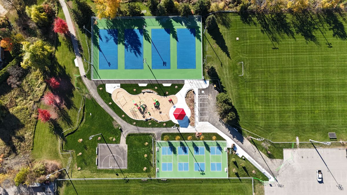 Aerial view of a park featuring tennis and pickleball courts, a half basketball court, a playground, and green fields.