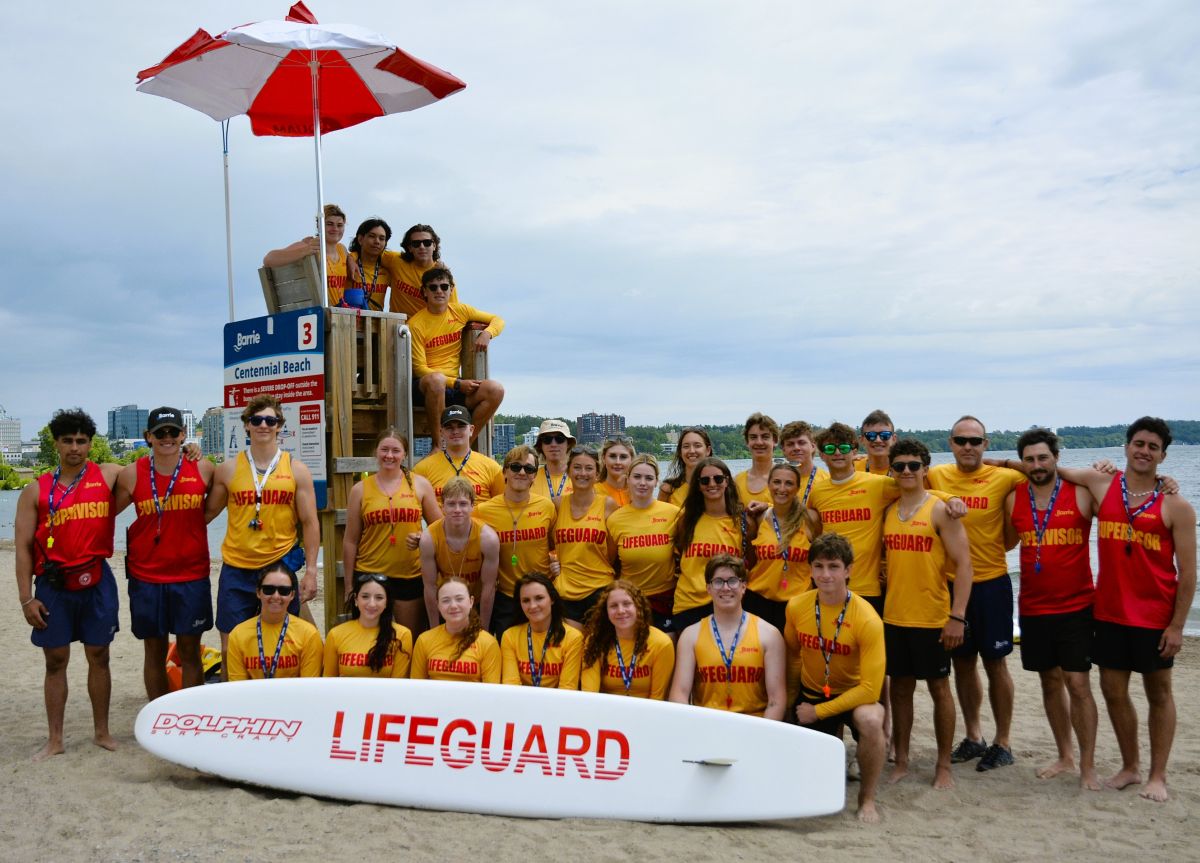 A group of lifeguards in yellow shirts poses on a beach by a lifeguard tower.