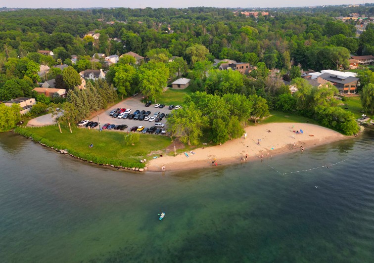 Aerial view of a sandy beach with green trees, parking lot, and people enjoying the water.