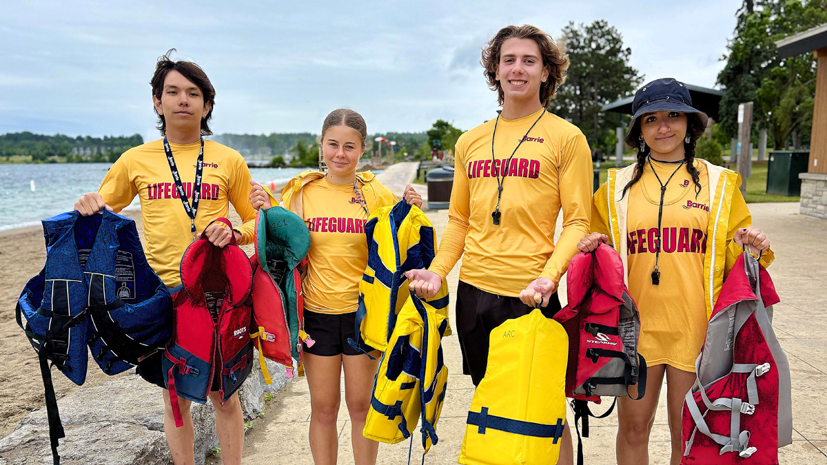 Four lifeguards in yellow shirts stand on a beach holding life vests next to the water.
