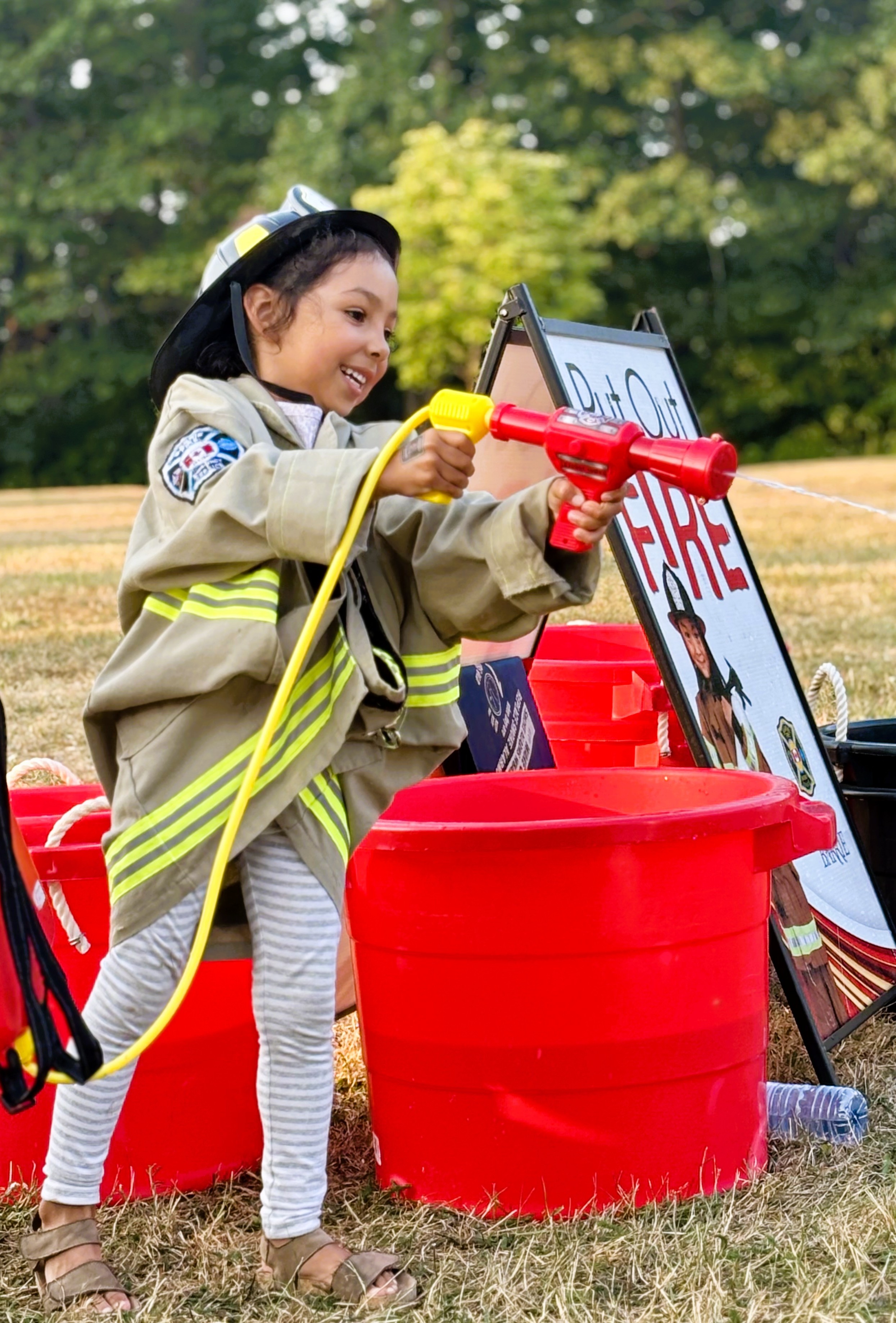 A young girl in a firefighter costume uses a toy fire hose, smiling and engaged in play.