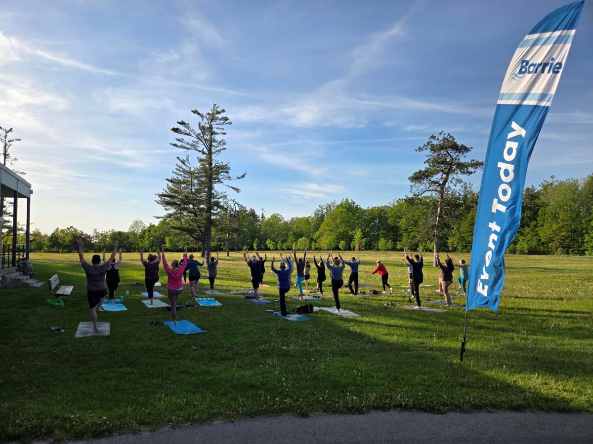 Group of people practicing yoga outdoors on mats beneath a clear blue sky. A Barrie "event today" banner is on display.