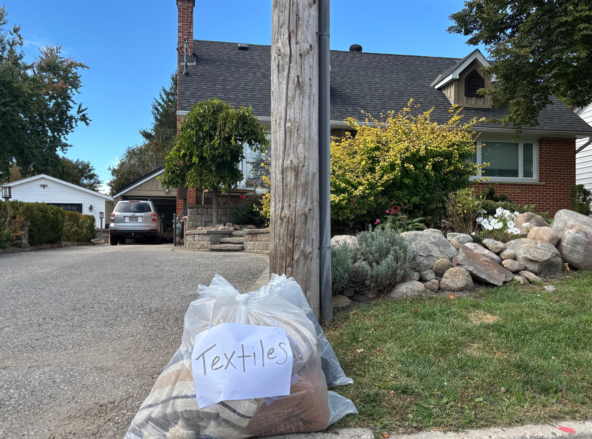 A bag labeled "Textiles" sits by a curb near a house and driveway.