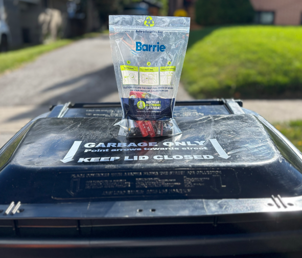 A clear bag with the Barrie logo filled with batteries on a black garbage cart outside, with grass in the background.