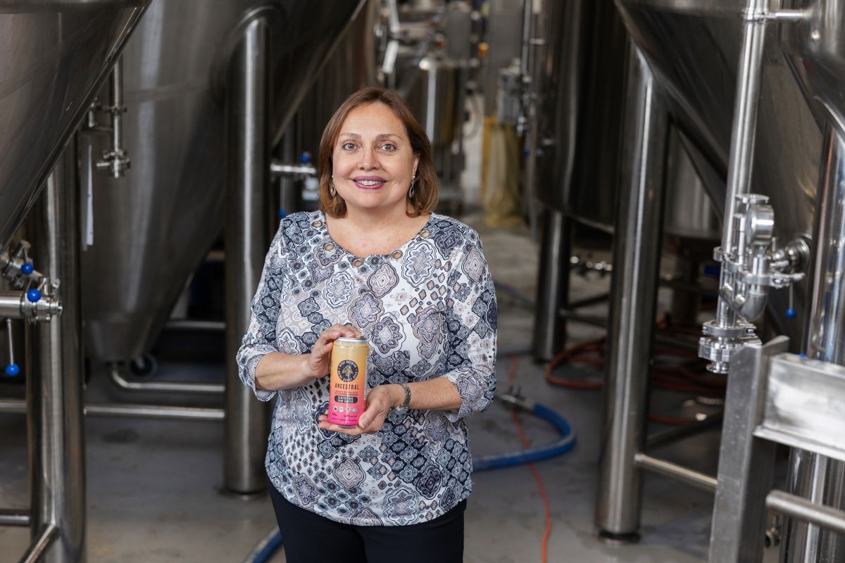 A person smiling and holding a beverage can stands in a brewery with large metal tanks.