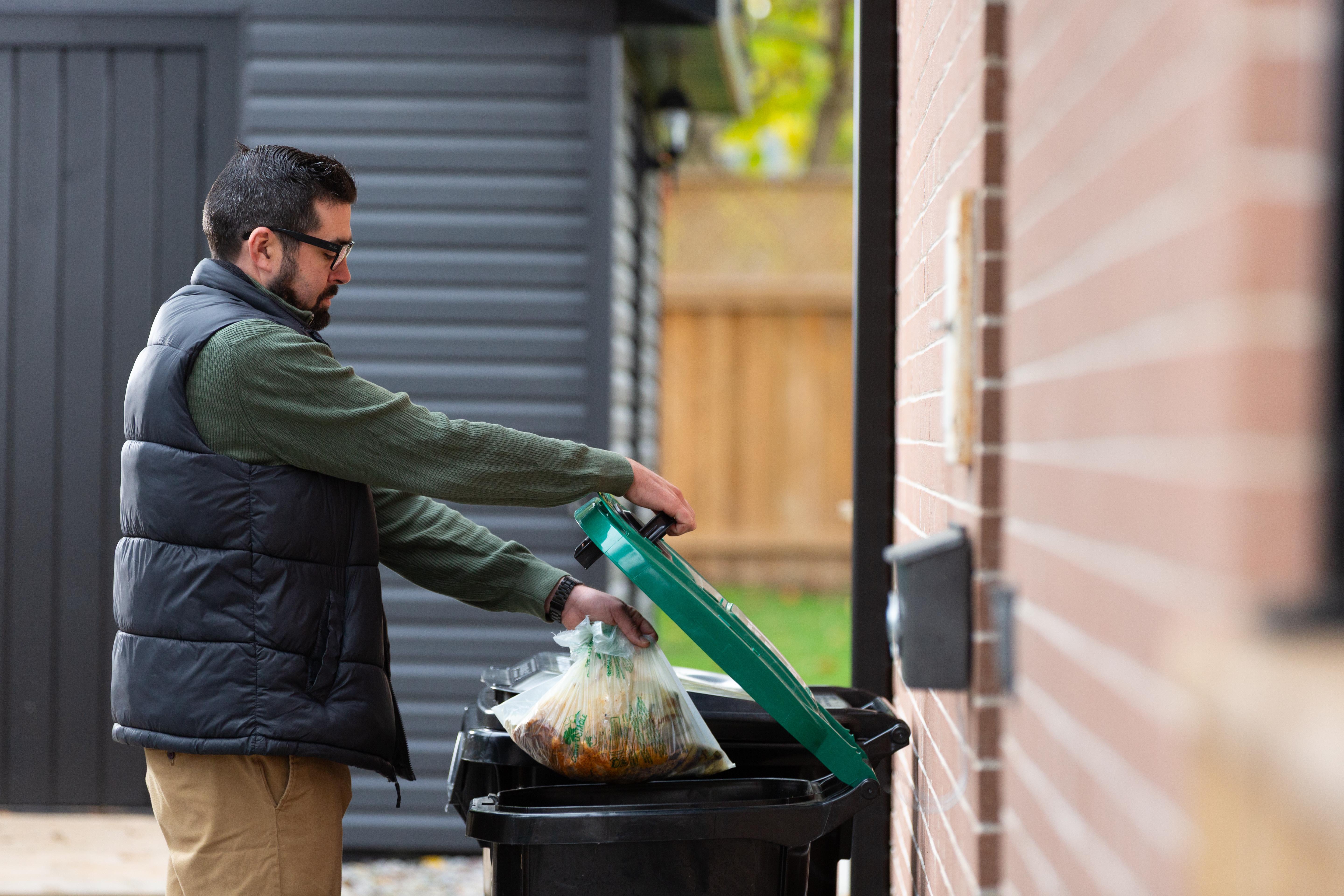 Person discarding a bag of organic waste into an organics cart beside a brick wall.