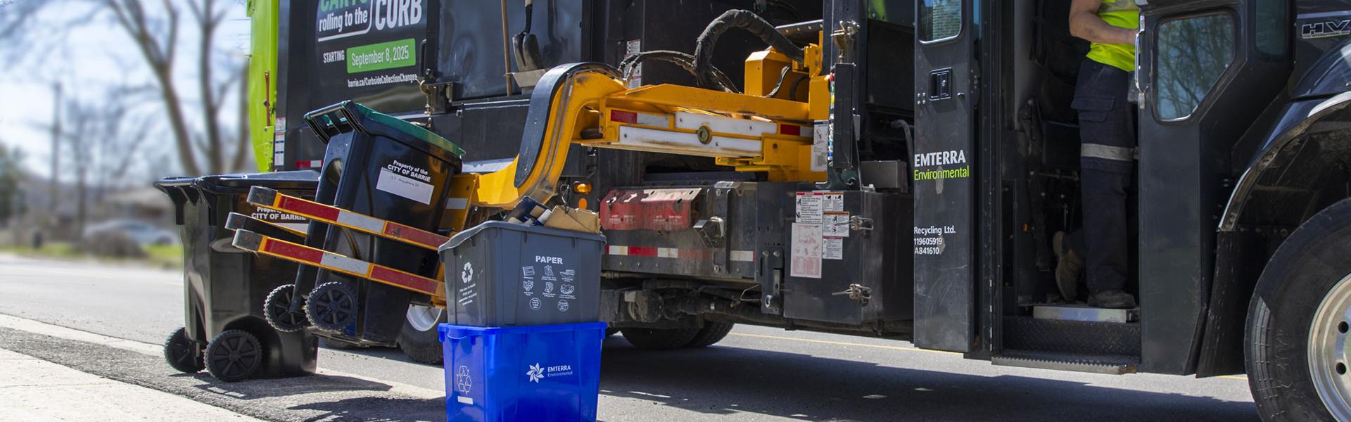 A waste management truck collects an organics cart from the curb, assisted by a worker in high visibility clothing.