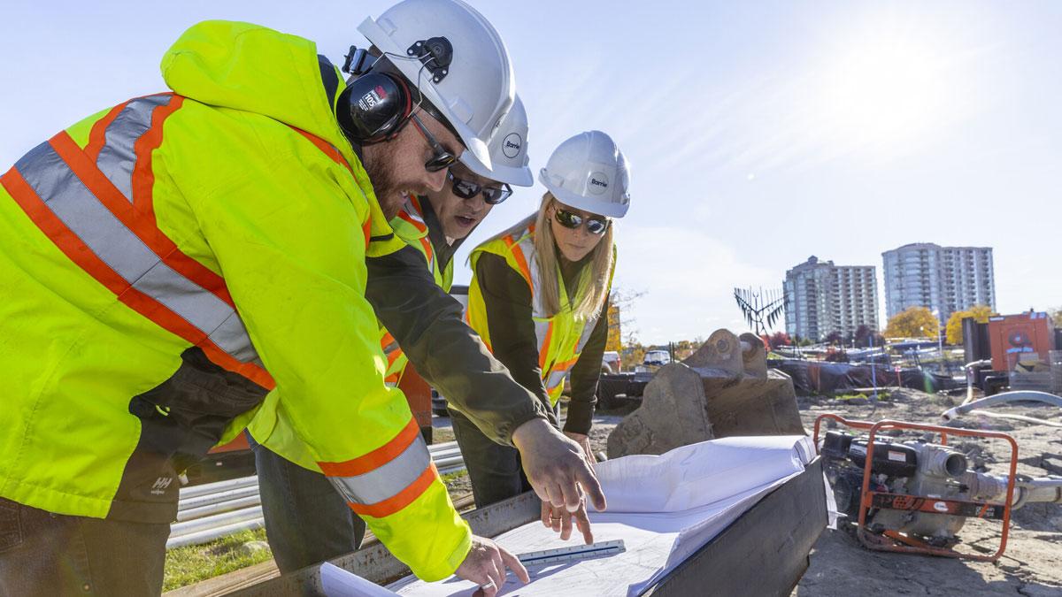 Three people with hard hats and safety vests looking at plans, bright sky and condo buildings in background