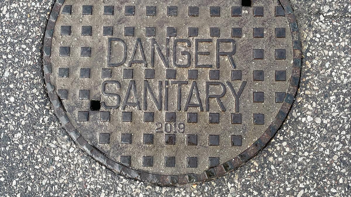 Manhole cover inscribed with the words 'DANGER SANITARY 2019' set in asphalt.