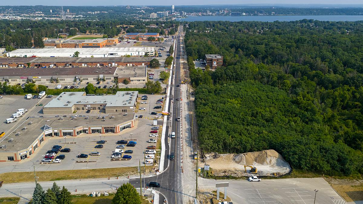 Aerial view featuring a road under construction and two-way traffic, flanked by commercial buildings on one side; Kempenfelt Bay and downtown Barrie in background.