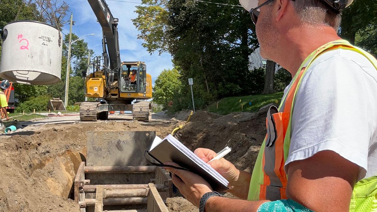 A construction worker in a high-visibility vest is observing and taking notes beside an excavation site where a large digger is operating near another worker.