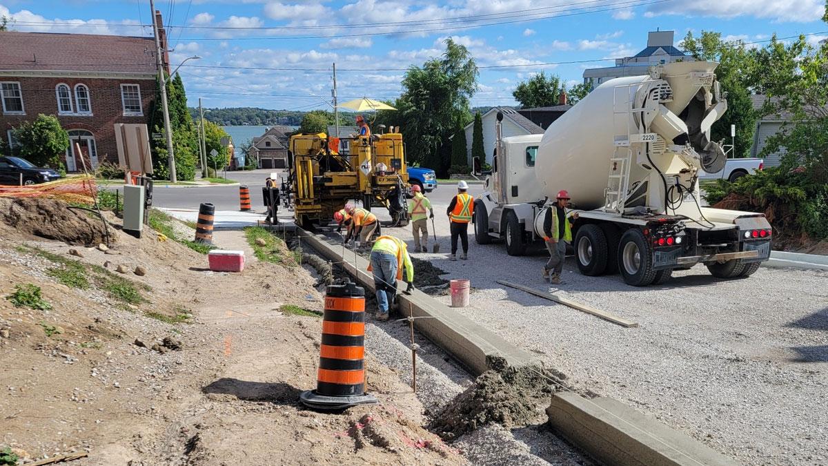 Workers operating machinery and performing road construction in a sunny, urban setting.