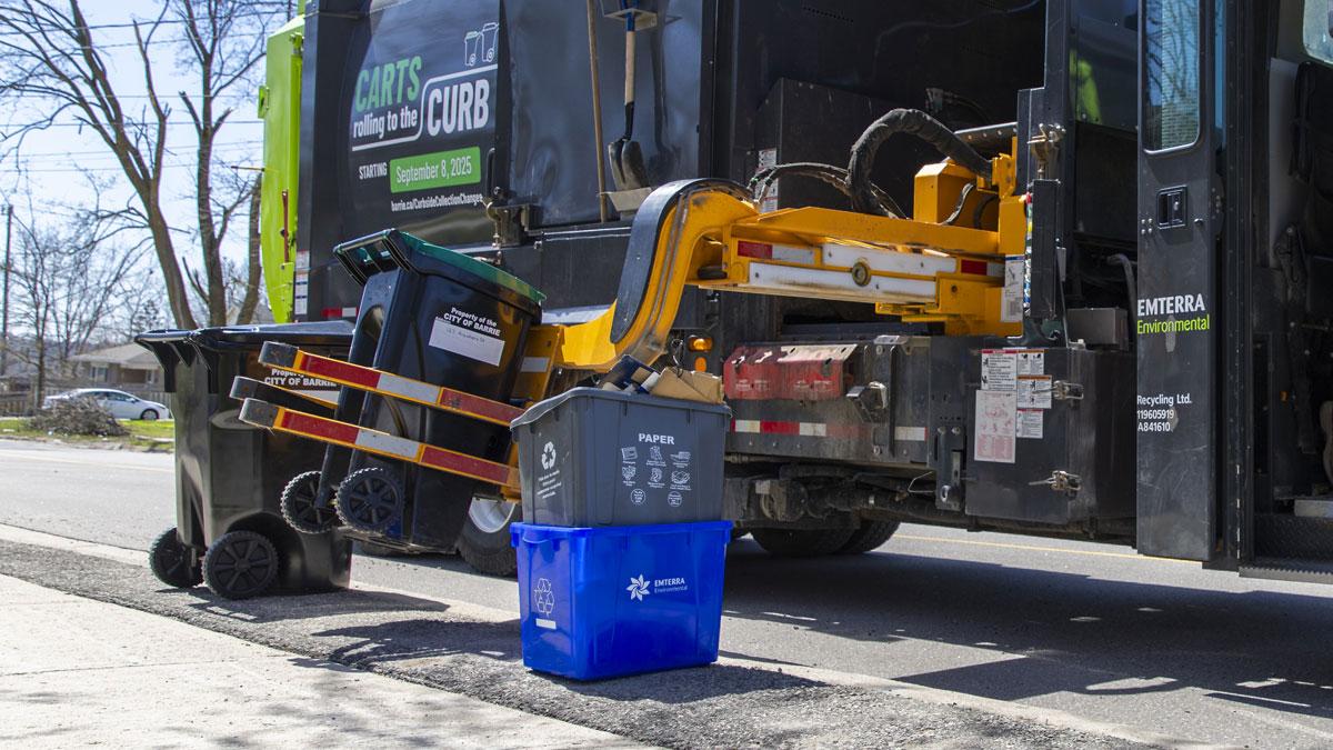 A waste management truck collects an organics cart from the curb, assisted by a worker in high visibility clothing.