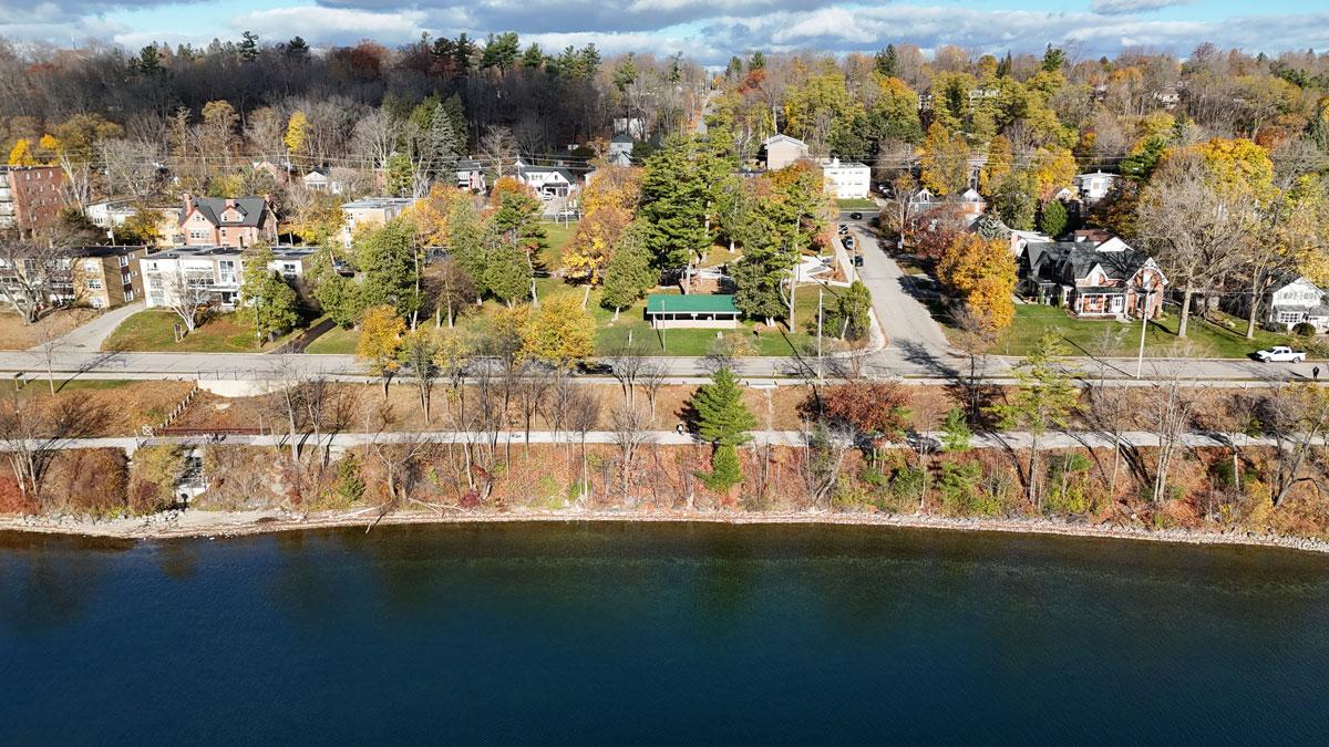 Aerial view of a trail and road beside a body of water, featuring residential buildings surrounded by trees with autumn foliage.