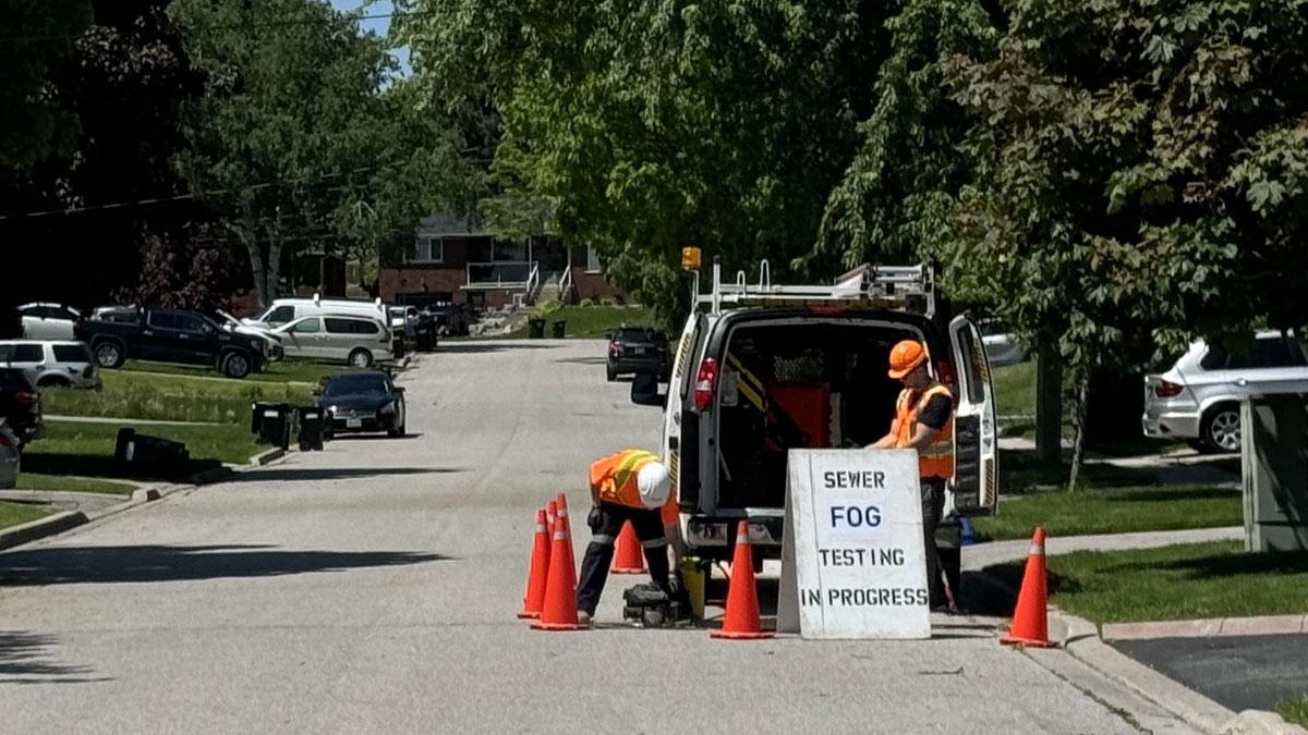 Workers in safety vests and hard hats conduct sewer fog testing on a residential street, with a sign stating "SEWER FOG TESTING IN PROGRESS" and orange traffic cones around a utility vehicle.