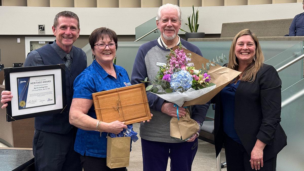 Four people smiling, holding a certificate, flowers, and a wooden plaque.