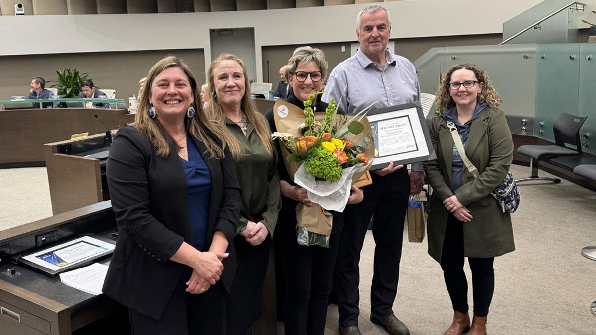 Group of five people smiling, one holds flowers and another holds a framed certificate.