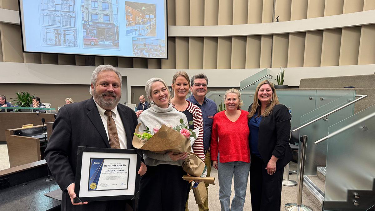 Group of smiling people, one holding a certificate, another with flowers.