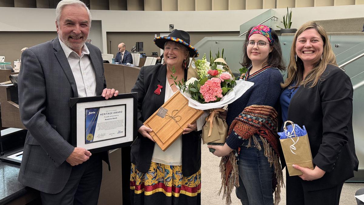Four people smiling, one holding a certificate, others with flowers and gift bags.