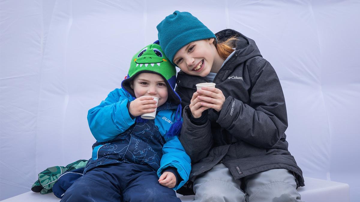 Two smiling children in winter clothing, holding cups.