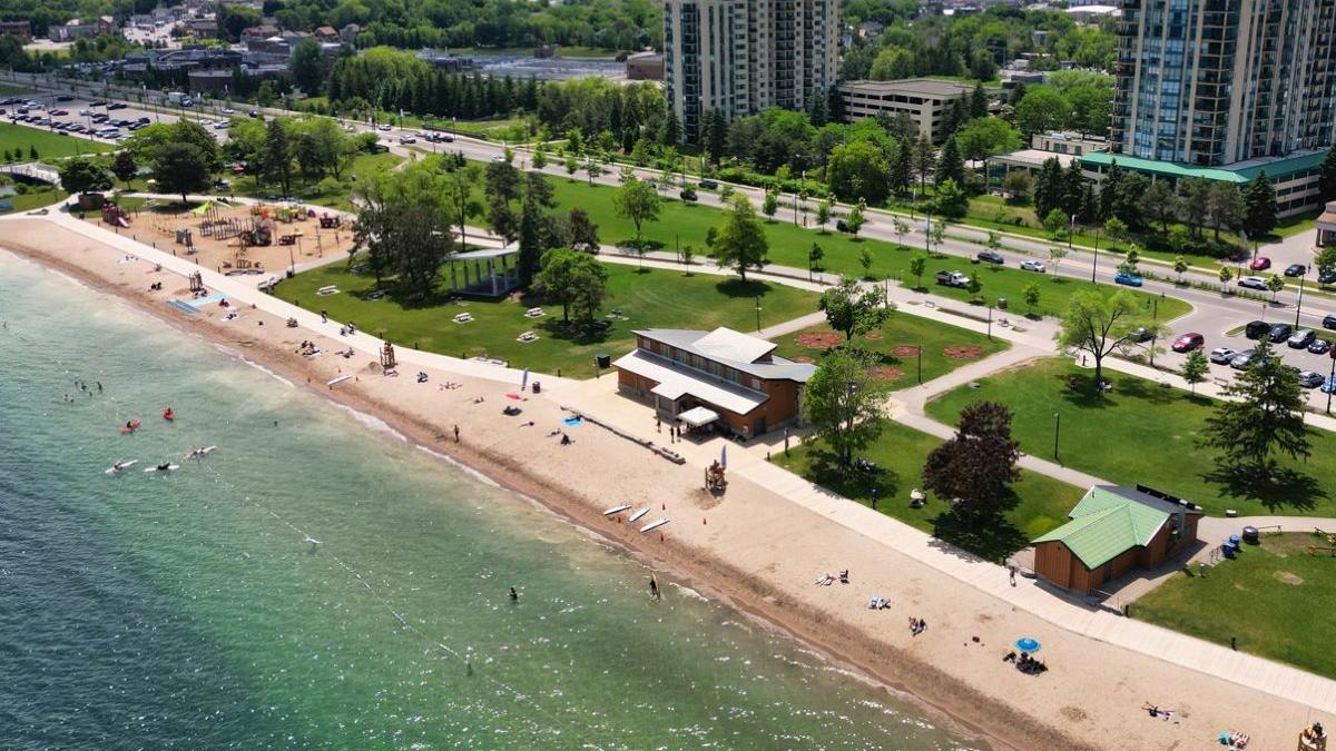 Aerial view of a beach with people, sandy shore, green parkland, and tall buildings in the background.