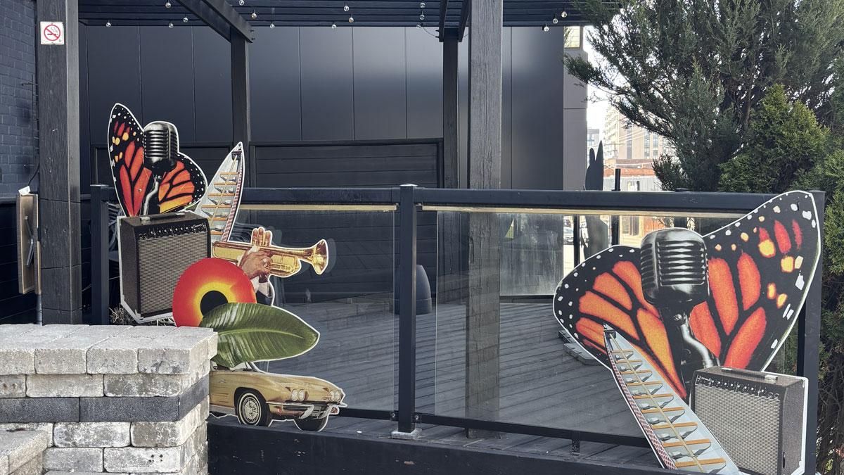 Colourful collage on a patio fence including a butterfly wing, microphone, speaker, vintage car, a brass instrument, canoe and more.