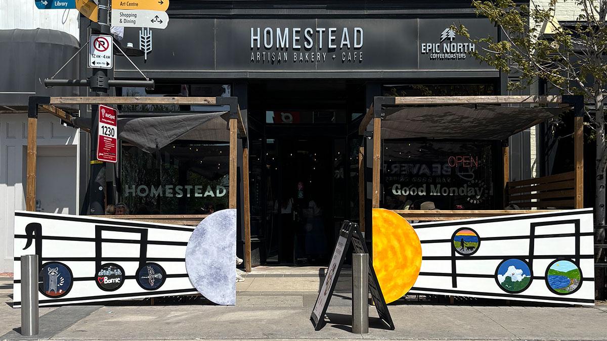 Exterior of Homestead Cafe with artwork on the patio fence of a moon and sun, and decorative musical notes featuring a racoon, the Heart Barrie sign, a bird, a person walking their dog on the waterfront, clouds and trees, and a body of water.