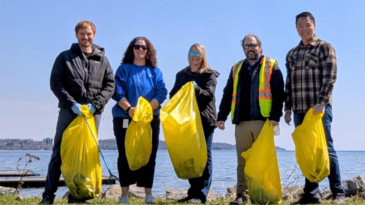 Five people holding yellow trash bags stand by a lake on a sunny day.