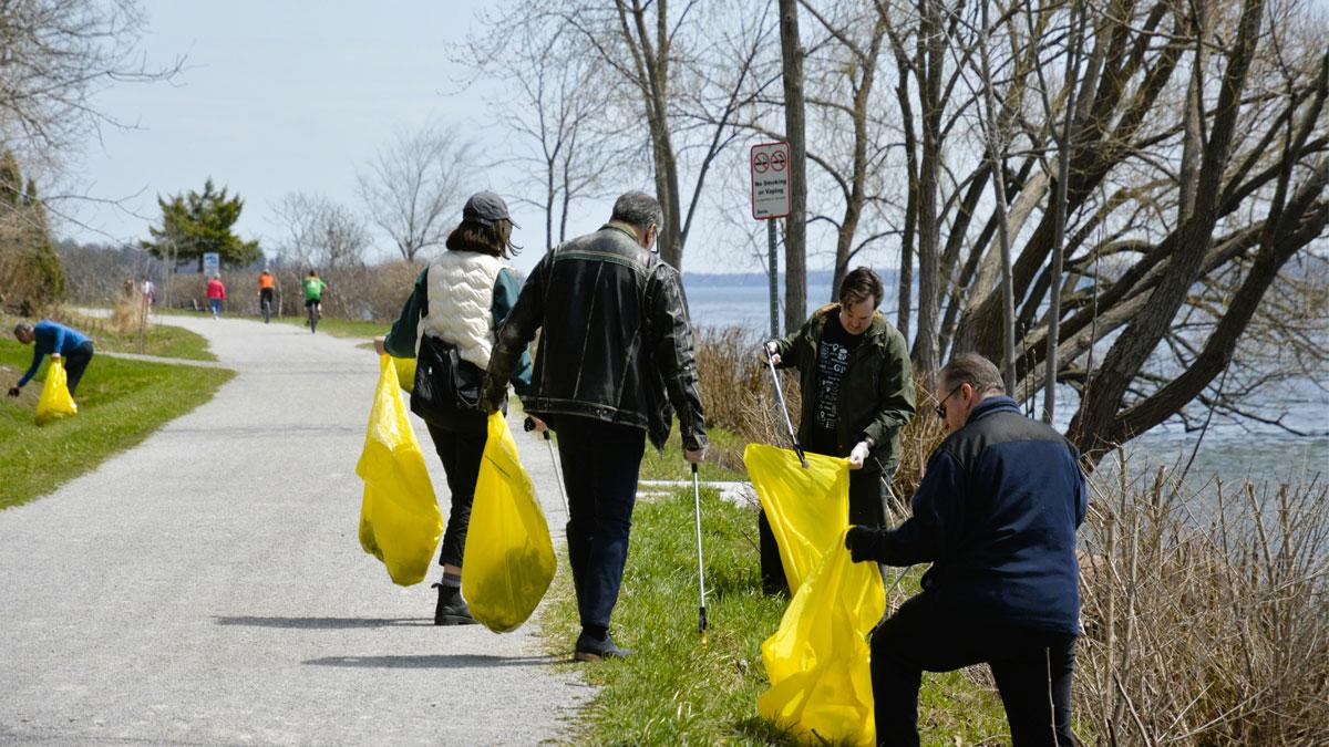 People collecting trash along a waterfront path on a sunny day, using yellow bags.