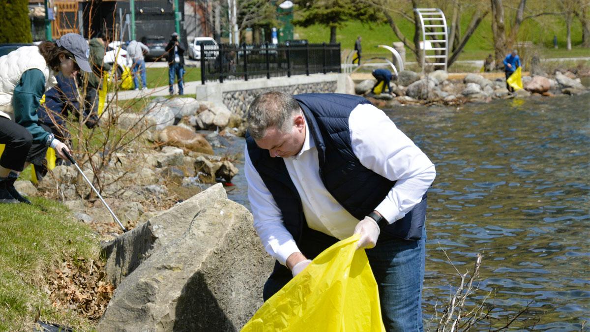 A man collects trash in a yellow bag by the water, with others cleaning in the background.