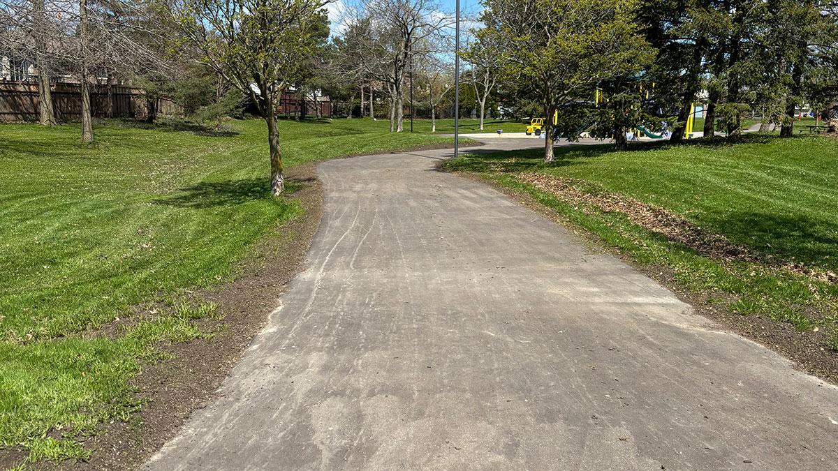 Winding pathway to a playground through a grassy area with trees under a clear blue sky.