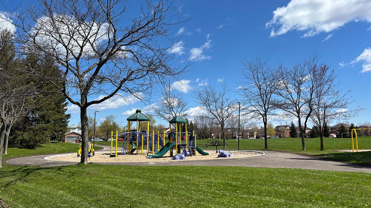 Sunny park scene featuring a playground surrounded by fresh pavement, green grass, and trees under a blue sky.