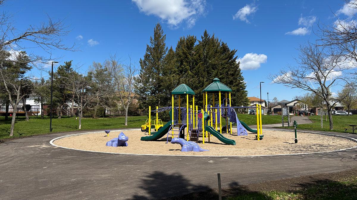 Colorful playground equipment surrounded by fresh pavement and a grassy area with sparse trees under a blue sky.