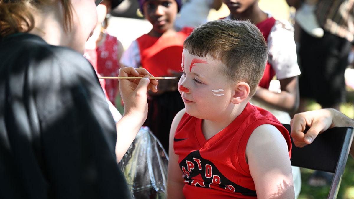 A young child in a red shirt has their face painted by a smiling artist outdoors.