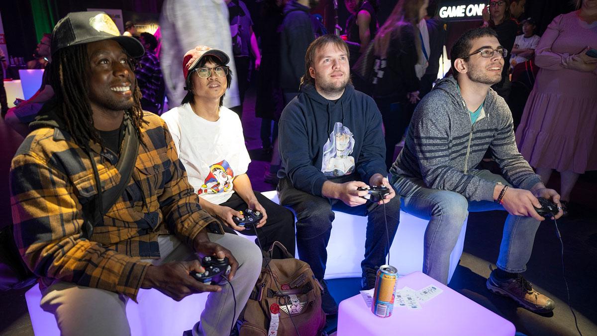 Four people are seated, playing video games, with colourful lighting and excitement in the background.
