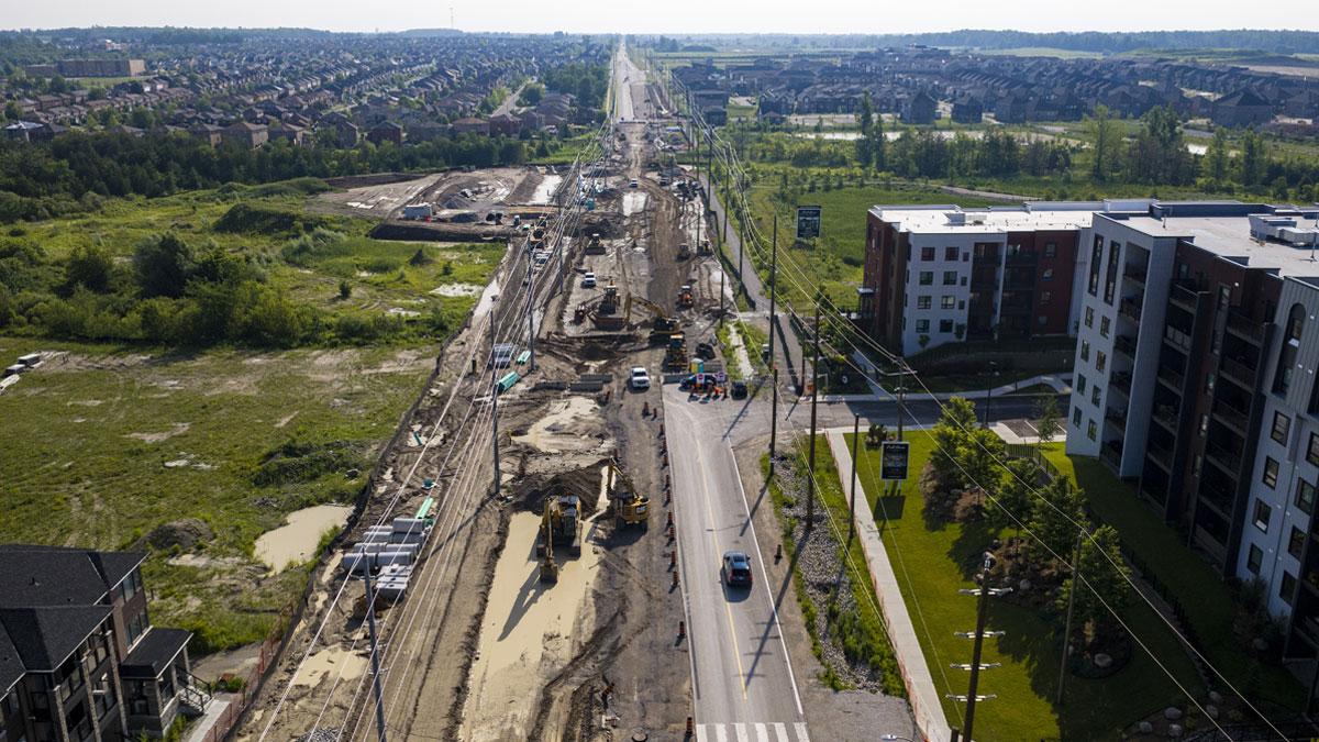 Aerial view of a road under construction, with buildings and green fields nearby.