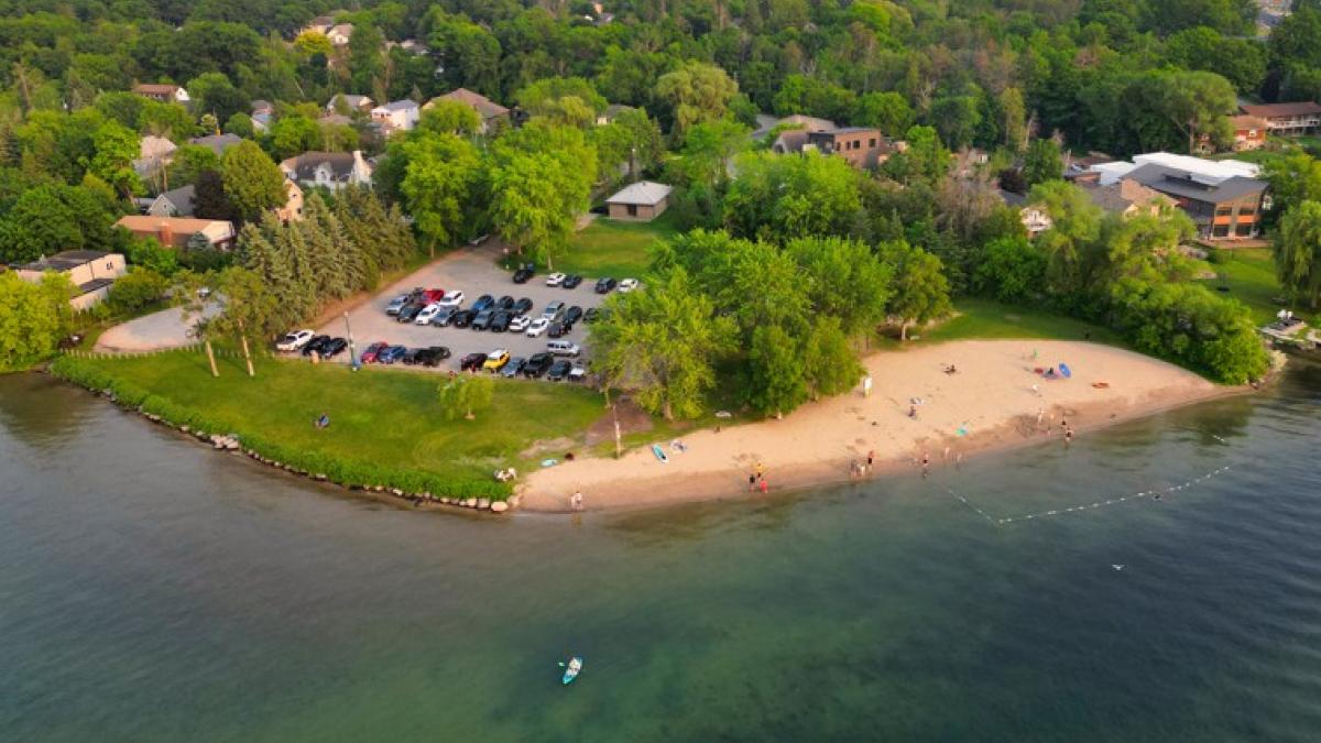 Aerial view of a sandy beach with green trees, parking lot, and people enjoying the water.