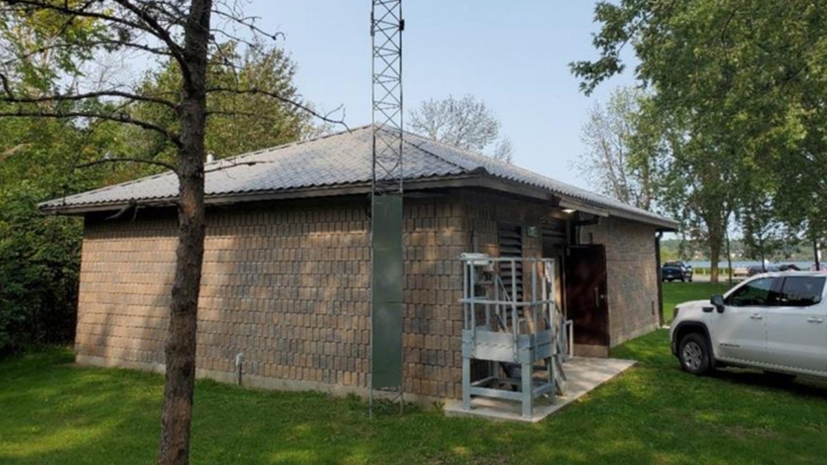 A small brick building with a metal tower and equipment outside, surrounded by grass and trees.