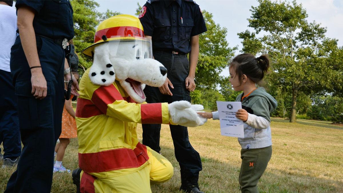 A costumed dog in a firefighter outfit kneels to interact with a smiling child in a park.