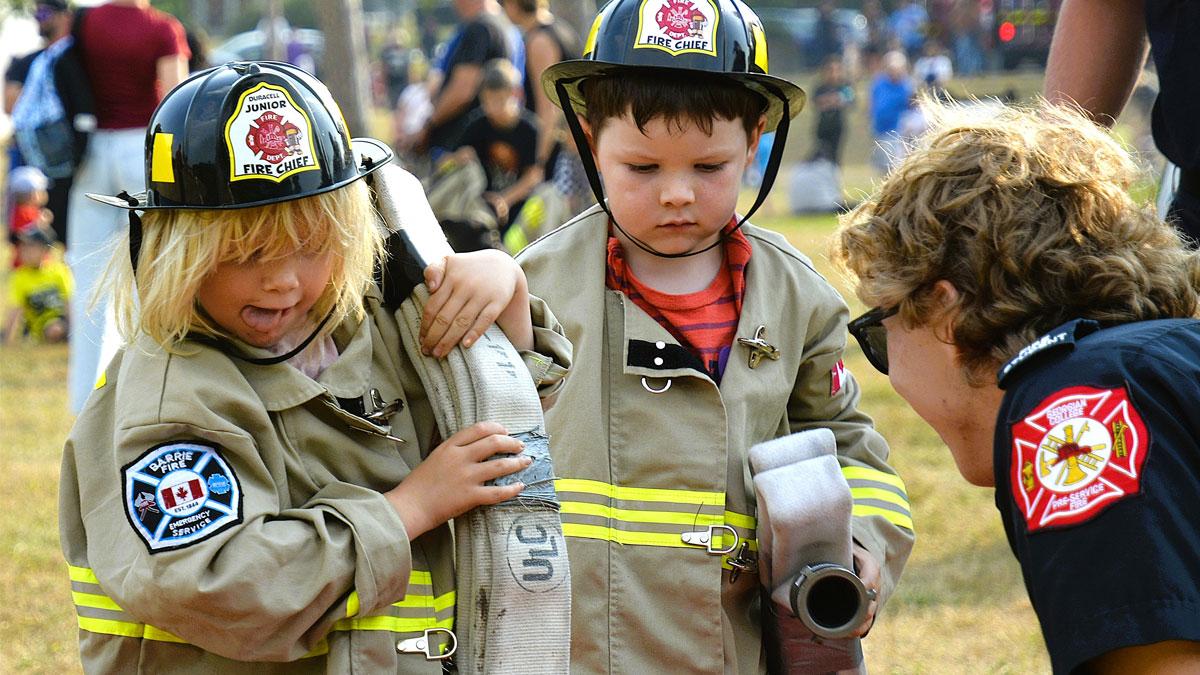 Two children in firefighter uniforms hold equipment while interacting with an adult.