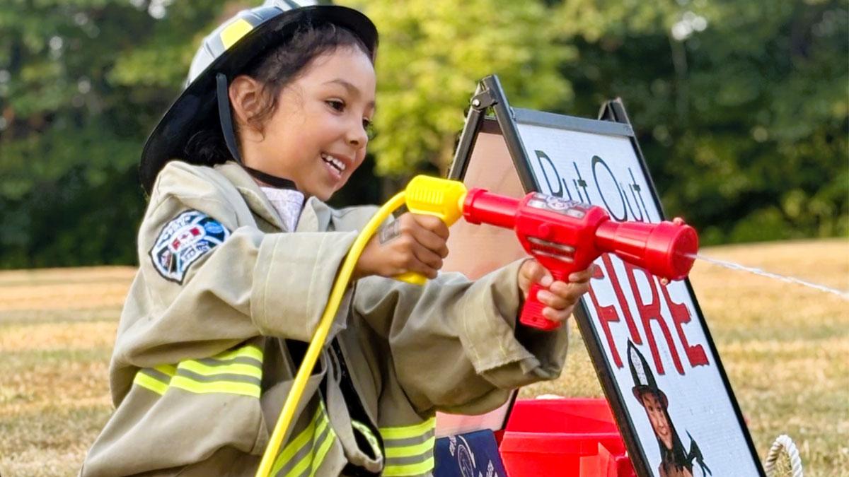 A child in a firefighter uniform playfully uses a water hose toy outdoors.