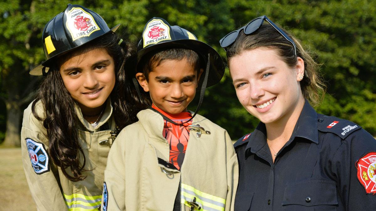 Two children wearing firefighter outfits and hats, pose with a smiling firefighter.